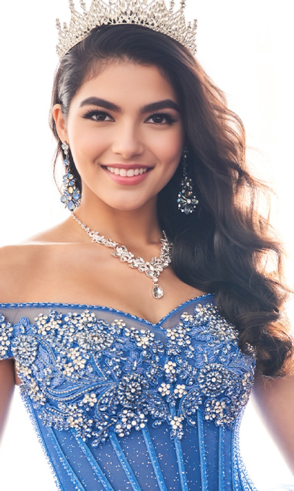 A woman smiles at the camera wearing the Quinceanera Dress PR30180 By Princesa, featuring an off-the-shoulder blue beaded gown, jeweled crown, and matching jewelry against a bright background.
